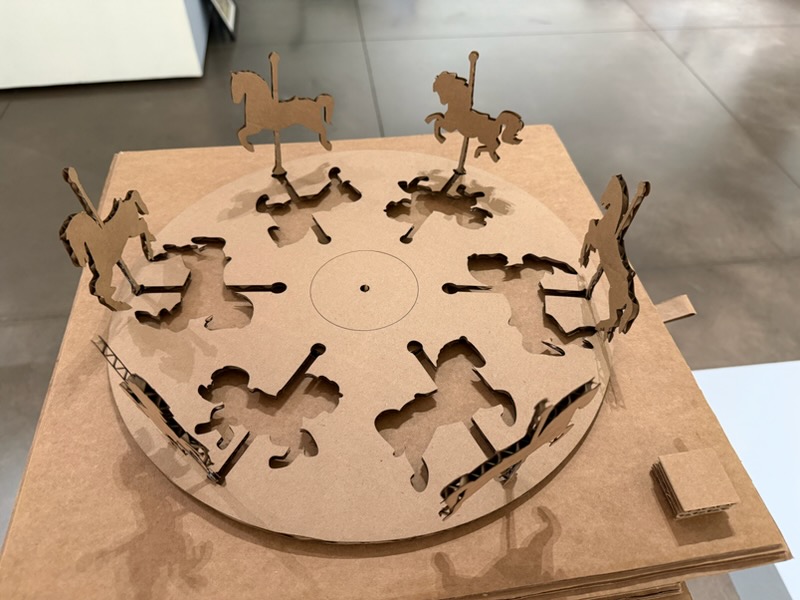 A laser-cut cardboard carousel sculpture with horse figures arranged in a circle on a rotating platform
