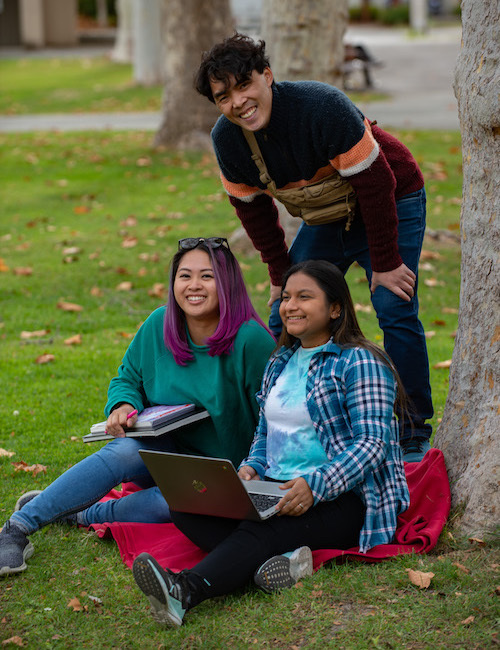 three students outside: one male standing, two women seated